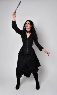 Siyah Viktorya dönemi korseli cadı kostümü giyen, stüdyo arka planında el hareketleriyle duran koyu renk saçlı kadının tam boy portresi..