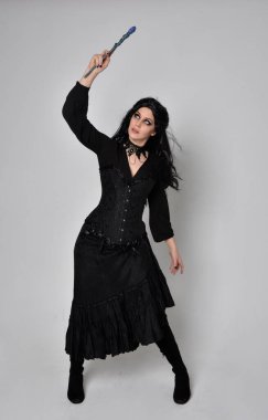 Siyah Viktorya dönemi korseli cadı kostümü giyen, stüdyo arka planında el hareketleriyle duran koyu renk saçlı kadının tam boy portresi..