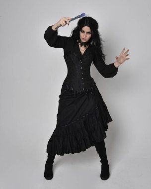 Full length portrait of dark haired woman wearing  black victorian witch costume with corset,  standing pose with  gestural hand movements,  against studio background. clipart