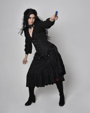 Siyah Viktorya dönemi korseli cadı kostümü giyen, stüdyo arka planında el hareketleriyle duran koyu renk saçlı kadının tam boy portresi..