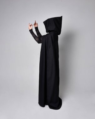 Siyah Victoria Cadı kostümü giymiş siyah saçlı, pelerinli ve sivri şapkalı bir kadının tam boy portresi. Ayakta duruş, arka plan, hareketli el hareketleri, stüdyo arka planı.