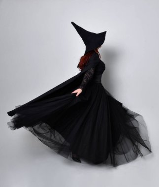 Siyah Victoria Cadı kostümü giymiş siyah saçlı, pelerinli ve sivri şapkalı bir kadının tam boy portresi. Ayakta duruş, arka plan, hareketli el hareketleri, stüdyo arka planı.
