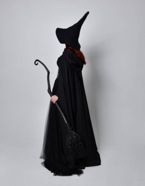 Siyah Victoria Cadı kostümü giymiş siyah saçlı, pelerinli ve sivri şapkalı bir kadının tam boy portresi. Ayakta duruş, arka plan, hareketli el hareketleri, stüdyo arka planı.