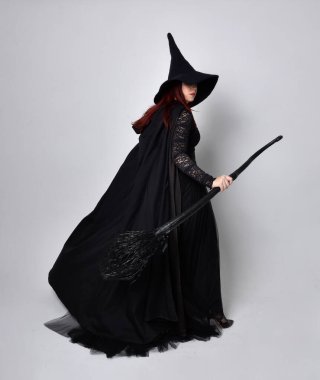 Siyah Victoria Cadı kostümü giymiş siyah saçlı, pelerinli ve sivri şapkalı bir kadının tam boy portresi. Ayakta duruş, arka plan, hareketli el hareketleri, stüdyo arka planı.