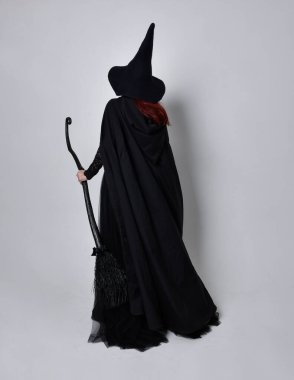 Siyah Victoria Cadı kostümü giymiş siyah saçlı, pelerinli ve sivri şapkalı bir kadının tam boy portresi. Ayakta duruş, arka plan, hareketli el hareketleri, stüdyo arka planı.