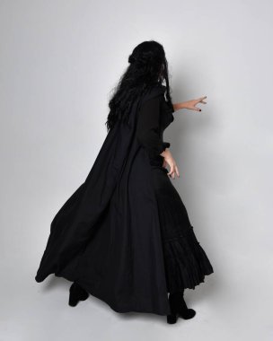 Siyah Victoria Cadı kostümü giymiş siyah saçlı, pelerinli ve sivri şapkalı bir kadının tam boy portresi. Ayakta duruş, arka plan, hareketli el hareketleri, stüdyo arka planı.