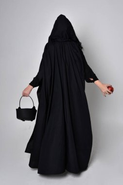 Siyah Victoria Cadı kostümü giymiş siyah saçlı, pelerinli ve sivri şapkalı bir kadının tam boy portresi. Ayakta duruş, arka plan, hareketli el hareketleri, stüdyo arka planı.