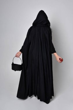 Siyah Victoria Cadı kostümü giymiş siyah saçlı, pelerinli ve sivri şapkalı bir kadının tam boy portresi. Ayakta duruş, arka plan, hareketli el hareketleri, stüdyo arka planı.