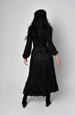 Siyah Victoria cadısı kostümü giymiş koyu renk saçlı bir kadının tam boy portresi. Ayakta duruş, arka plan, hareketli el hareketleri, stüdyo arka planı.