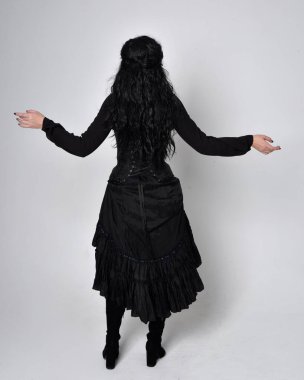 Siyah Victoria cadısı kostümü giymiş koyu renk saçlı bir kadının tam boy portresi. Ayakta duruş, arka plan, hareketli el hareketleri, stüdyo arka planı.