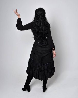 Siyah Victoria cadısı kostümü giymiş koyu renk saçlı bir kadının tam boy portresi. Ayakta duruş, arka plan, hareketli el hareketleri, stüdyo arka planı.