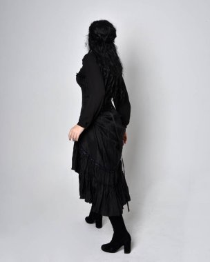 Siyah Victoria cadısı kostümü giymiş koyu renk saçlı bir kadının tam boy portresi. Ayakta duruş, arka plan, hareketli el hareketleri, stüdyo arka planı.