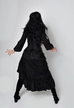 Siyah Victoria cadısı kostümü giymiş koyu renk saçlı bir kadının tam boy portresi. Ayakta duruş, arka plan, hareketli el hareketleri, stüdyo arka planı.