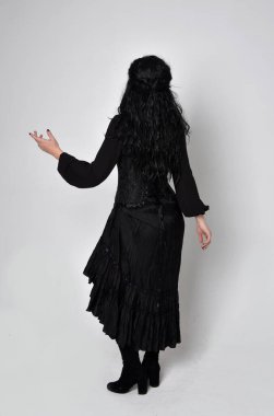 Siyah Victoria cadısı kostümü giymiş koyu renk saçlı bir kadının tam boy portresi. Ayakta duruş, arka plan, hareketli el hareketleri, stüdyo arka planı.