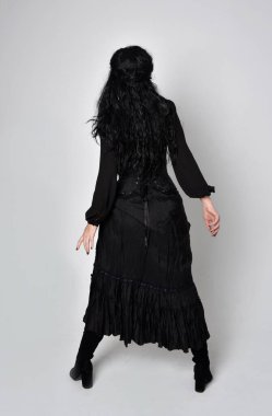 Siyah Victoria cadısı kostümü giymiş koyu renk saçlı bir kadının tam boy portresi. Ayakta duruş, arka plan, hareketli el hareketleri, stüdyo arka planı.