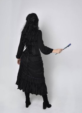 Siyah Victoria cadısı kostümü giymiş koyu renk saçlı bir kadının tam boy portresi. Ayakta duruş, arka plan, hareketli el hareketleri, stüdyo arka planı.
