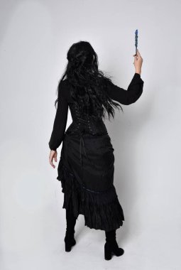 Siyah Victoria cadısı kostümü giymiş koyu renk saçlı bir kadının tam boy portresi. Ayakta duruş, arka plan, hareketli el hareketleri, stüdyo arka planı.