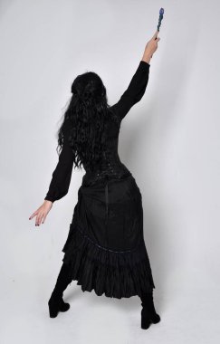 Siyah Victoria cadısı kostümü giymiş koyu renk saçlı bir kadının tam boy portresi. Ayakta duruş, arka plan, hareketli el hareketleri, stüdyo arka planı.