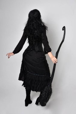 Siyah Victoria cadısı kostümü giymiş koyu renk saçlı bir kadının tam boy portresi. Ayakta duruş, arka plan, hareketli el hareketleri, stüdyo arka planı.