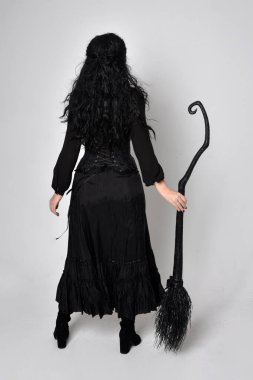 Siyah Victoria cadısı kostümü giymiş koyu renk saçlı bir kadının tam boy portresi. Ayakta duruş, arka plan, hareketli el hareketleri, stüdyo arka planı.