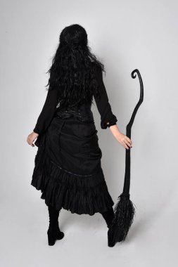 Siyah Victoria cadısı kostümü giymiş koyu renk saçlı bir kadının tam boy portresi. Ayakta duruş, arka plan, hareketli el hareketleri, stüdyo arka planı.