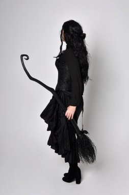 Siyah Victoria cadısı kostümü giymiş koyu renk saçlı bir kadının tam boy portresi. Ayakta duruş, arka plan, hareketli el hareketleri, stüdyo arka planı.