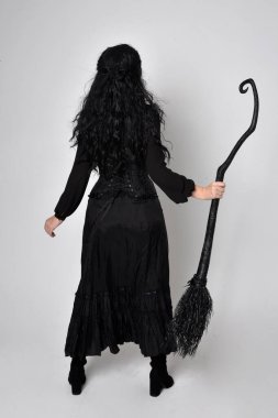 Siyah Victoria cadısı kostümü giymiş koyu renk saçlı bir kadının tam boy portresi. Ayakta duruş, arka plan, hareketli el hareketleri, stüdyo arka planı.