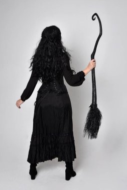 Siyah Victoria cadısı kostümü giymiş koyu renk saçlı bir kadının tam boy portresi. Ayakta duruş, arka plan, hareketli el hareketleri, stüdyo arka planı.