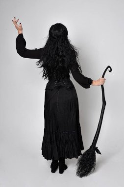 Siyah Victoria cadısı kostümü giymiş koyu renk saçlı bir kadının tam boy portresi. Ayakta duruş, arka plan, hareketli el hareketleri, stüdyo arka planı.