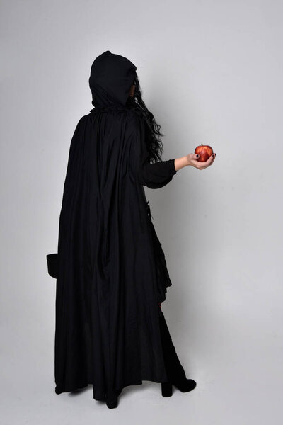 Полноразмерный портрет темноволосой женщины в чёрном викторианском костюме ведьмы в плаще и остроконечной шляпе. Стоя поза, вид сзади, с жестом движения рукой, на фоне студии.