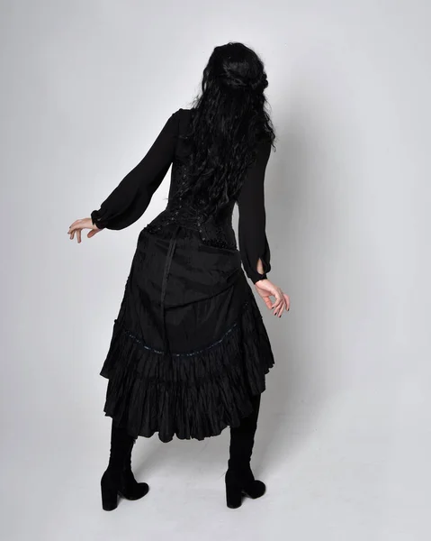 身穿黑色维多利亚式女巫服装的黑头发女人的全长肖像 倒立姿势 后视镜 有手势式的手部动作 在工作室的背景下 — 图库照片