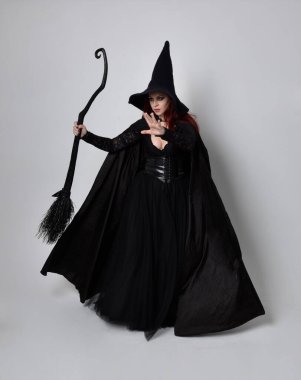 Siyah Victoria cadısı kostümü giymiş koyu renk saçlı bir kadının tam boy portresi. Stüdyonun arka planında el hareketleriyle bir süpürge sopası tutuyor..