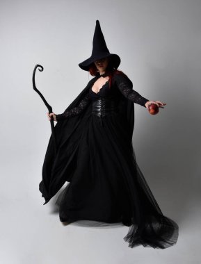 Siyah Victoria cadısı kostümü giymiş koyu renk saçlı bir kadının tam boy portresi. Stüdyonun arka planında el hareketleriyle bir süpürge sopası tutuyor..