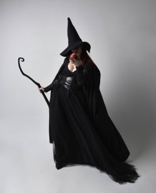 Siyah Victoria cadısı kostümü giymiş koyu renk saçlı bir kadının tam boy portresi. Stüdyonun arka planında el hareketleriyle bir süpürge sopası tutuyor..