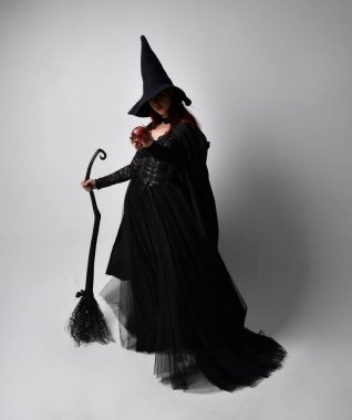 Siyah Victoria cadısı kostümü giymiş koyu renk saçlı bir kadının tam boy portresi. Stüdyonun arka planında el hareketleriyle bir süpürge sopası tutuyor..