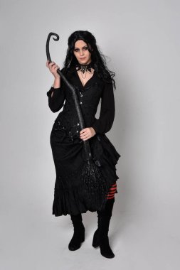 Siyah Victoria cadısı kostümü giymiş koyu renk saçlı bir kadının tam boy portresi. Stüdyonun arka planında el hareketleriyle bir süpürge sopası tutuyor..