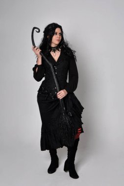 Siyah Victoria cadısı kostümü giymiş koyu renk saçlı bir kadının tam boy portresi. Stüdyonun arka planında el hareketleriyle bir süpürge sopası tutuyor..