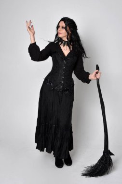 Siyah Victoria cadısı kostümü giymiş koyu renk saçlı bir kadının tam boy portresi. Stüdyonun arka planında el hareketleriyle bir süpürge sopası tutuyor..