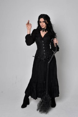 Siyah Victoria cadısı kostümü giymiş koyu renk saçlı bir kadının tam boy portresi. Stüdyonun arka planında el hareketleriyle bir süpürge sopası tutuyor..
