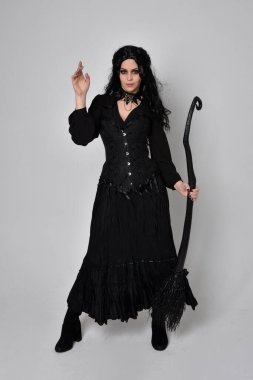 Siyah Victoria cadısı kostümü giymiş koyu renk saçlı bir kadının tam boy portresi. Stüdyonun arka planında el hareketleriyle bir süpürge sopası tutuyor..