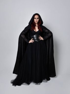 Siyah Viktorya dönemi cadı kostümü giymiş, pelerinli koyu renk saçlı bir kadının tam boy portresi. Stüdyonun arka planında, hareketli el hareketleri ile ayakta duruyoruz..