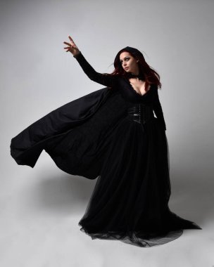Siyah Viktorya dönemi cadı kostümü giymiş, pelerinli koyu renk saçlı bir kadının tam boy portresi. Stüdyonun arka planında, hareketli el hareketleri ile ayakta duruyoruz..