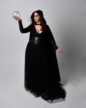 Full length portrait of dark haired woman wearing  black victorian witch costume with a flowing  cloak.  standing pose, with  gestural hand movements,  against studio background. clipart
