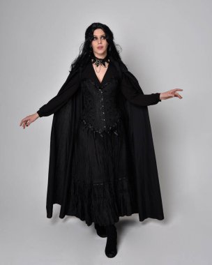 Siyah Viktorya dönemi cadı kostümü giymiş, pelerinli koyu renk saçlı bir kadının tam boy portresi. Stüdyonun arka planında, hareketli el hareketleri ile ayakta duruyoruz..