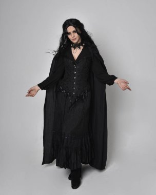 Siyah Viktorya dönemi cadı kostümü giymiş, pelerinli koyu renk saçlı bir kadının tam boy portresi. Stüdyonun arka planında, hareketli el hareketleri ile ayakta duruyoruz..