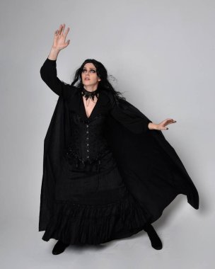 Siyah Viktorya dönemi cadı kostümü giymiş, pelerinli koyu renk saçlı bir kadının tam boy portresi. Stüdyonun arka planında, hareketli el hareketleri ile ayakta duruyoruz..