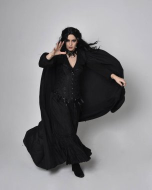 Siyah Viktorya dönemi cadı kostümü giymiş, pelerinli koyu renk saçlı bir kadının tam boy portresi. Stüdyonun arka planında, hareketli el hareketleri ile ayakta duruyoruz..