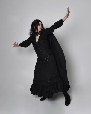 Siyah Viktorya dönemi cadı kostümü giymiş, pelerinli koyu renk saçlı bir kadının tam boy portresi. Stüdyonun arka planında, hareketli el hareketleri ile ayakta duruyoruz..