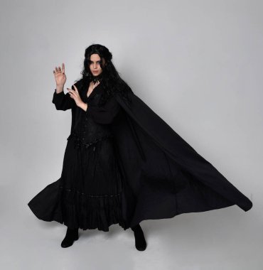 Siyah Viktorya dönemi cadı kostümü giymiş, pelerinli koyu renk saçlı bir kadının tam boy portresi. Stüdyonun arka planında, hareketli el hareketleri ile ayakta duruyoruz..