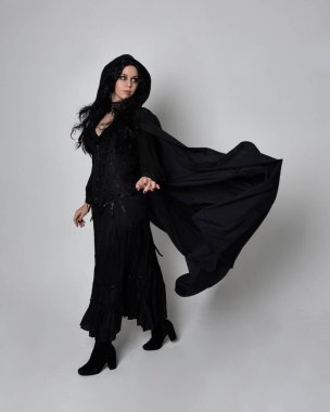 Siyah Viktorya dönemi cadı kostümü giymiş, pelerinli koyu renk saçlı bir kadının tam boy portresi. Stüdyonun arka planında, hareketli el hareketleri ile ayakta duruyoruz..